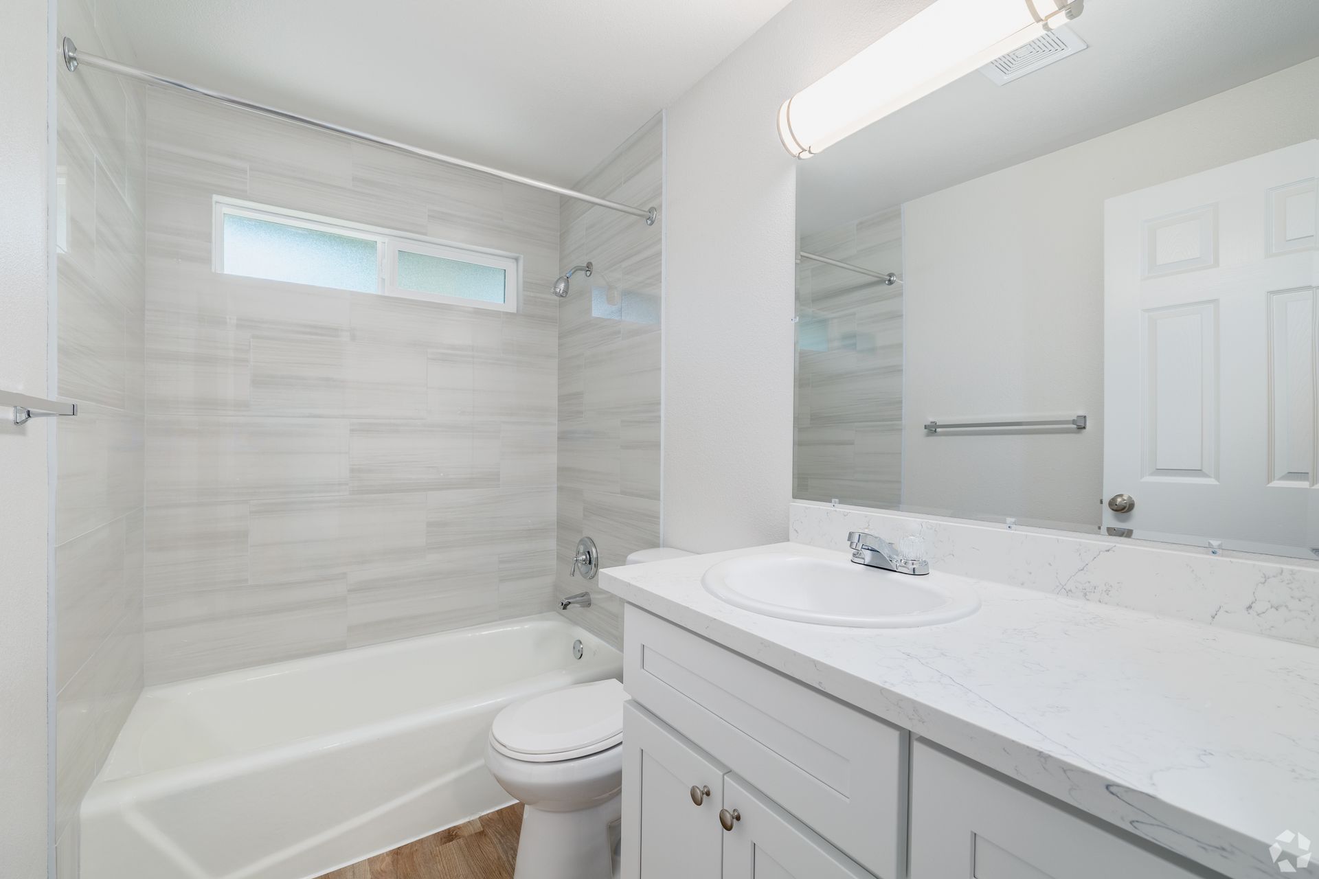 Bright white bathroom with a tub-shower combo, vanity, toilet, and small frosted window.