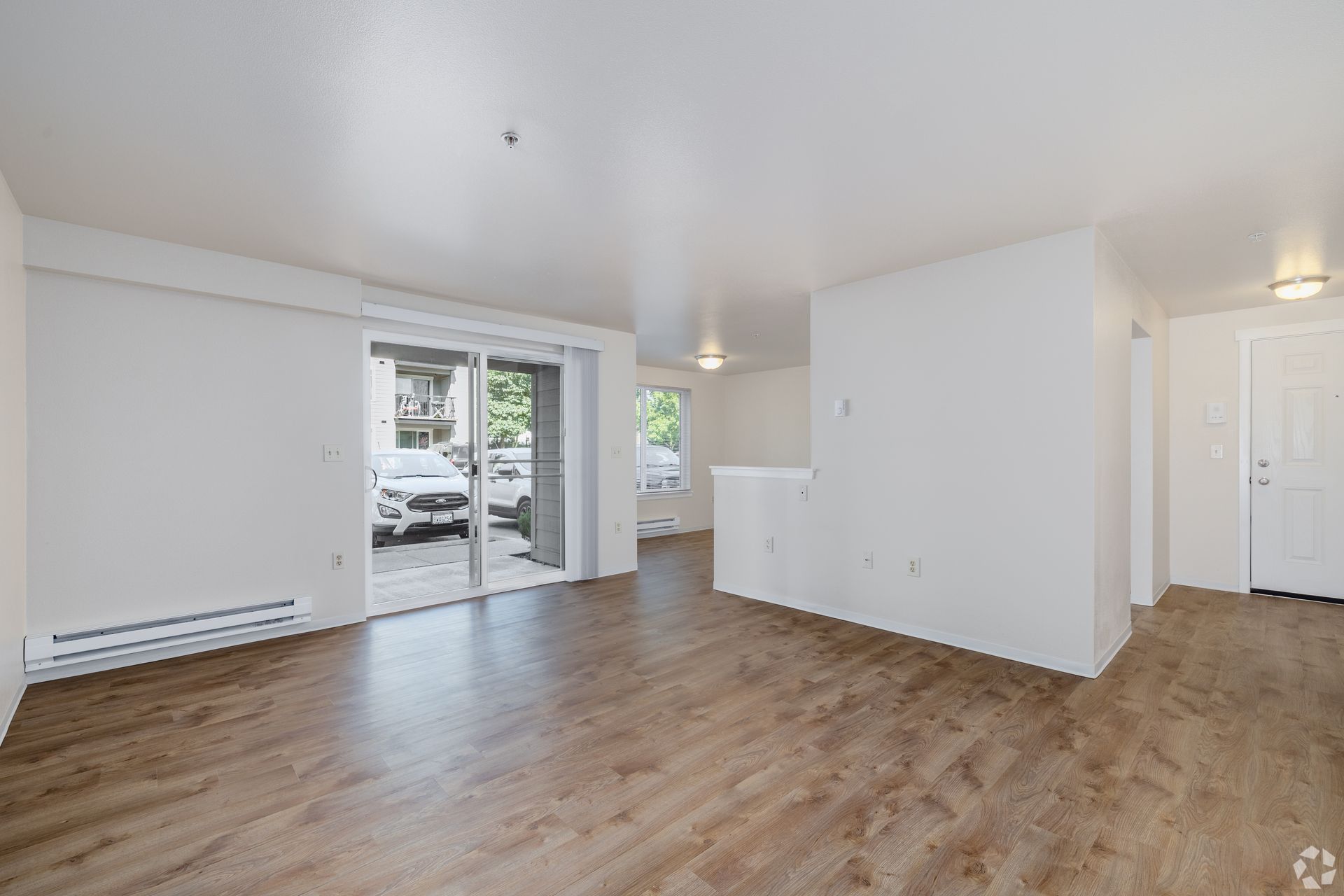 Empty living room with wood floors, white walls, sliding glass door, and hallway to the right.