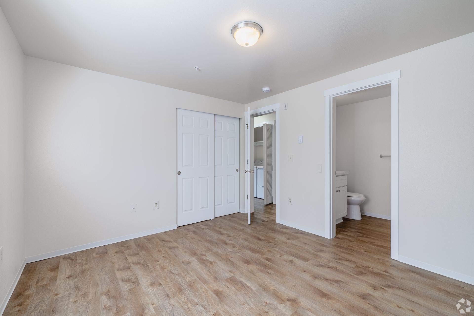 Empty white bedroom with wood floors, closet doors, and an open bathroom doorway