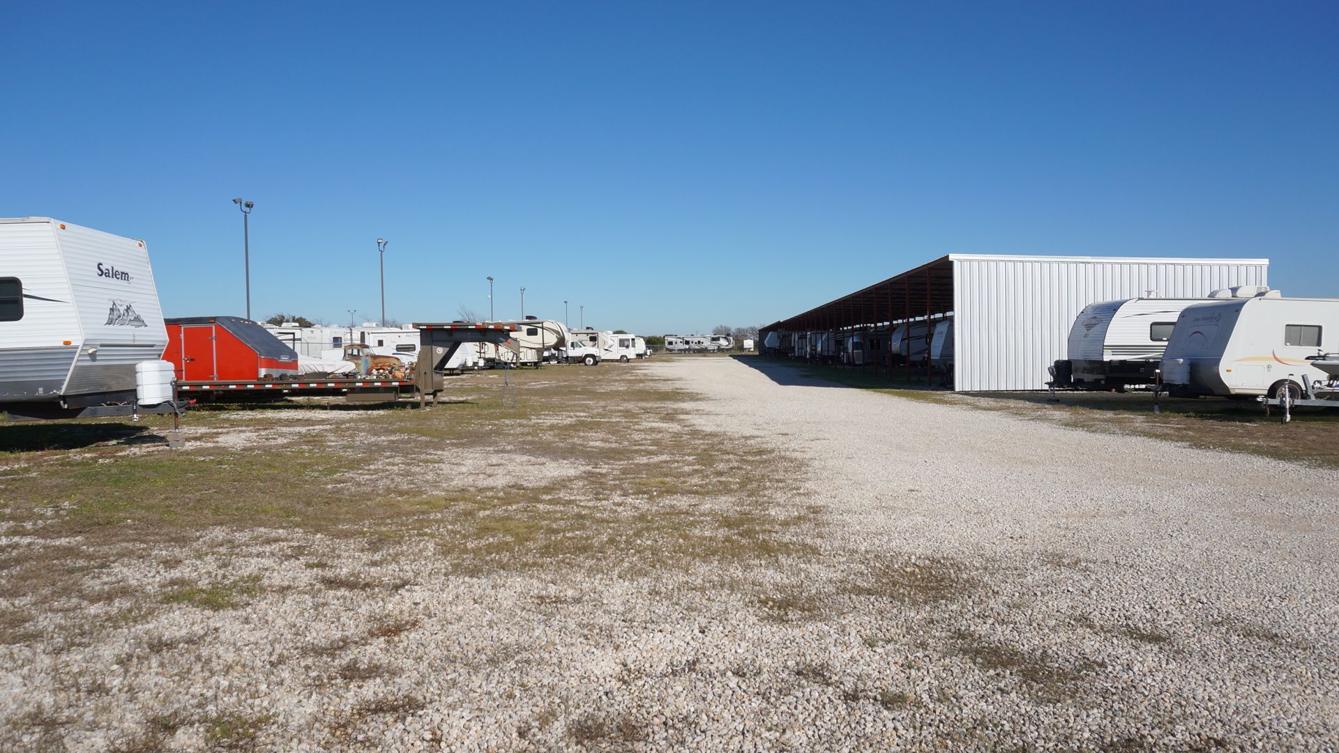 RVs parked in a gravel lot with a long, covered structure under a clear blue sky.