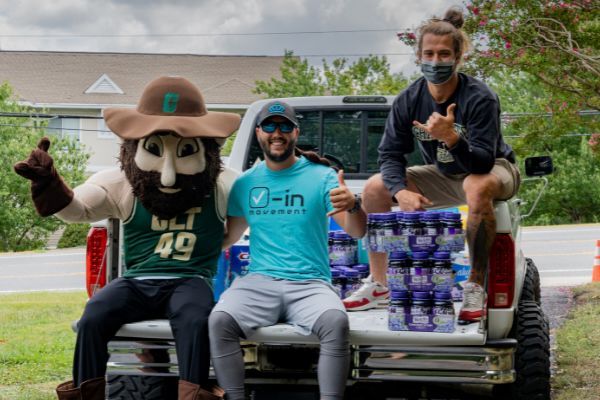 Three people posing on a pickup truck with stacked bottled water and a mascot in a parking lot