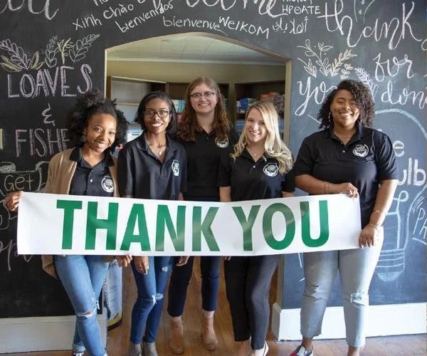 Five people hold a large green and white “THANK YOU” banner in front of a chalkboard wall.