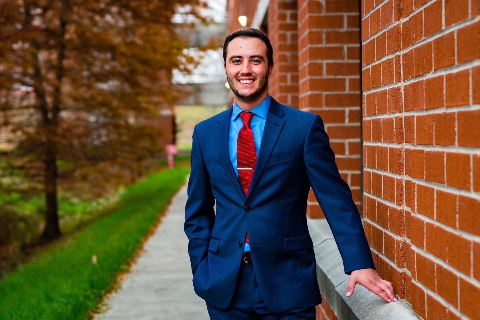 Man in a blue suit and red tie standing by a brick wall on a sidewalk lined with trees