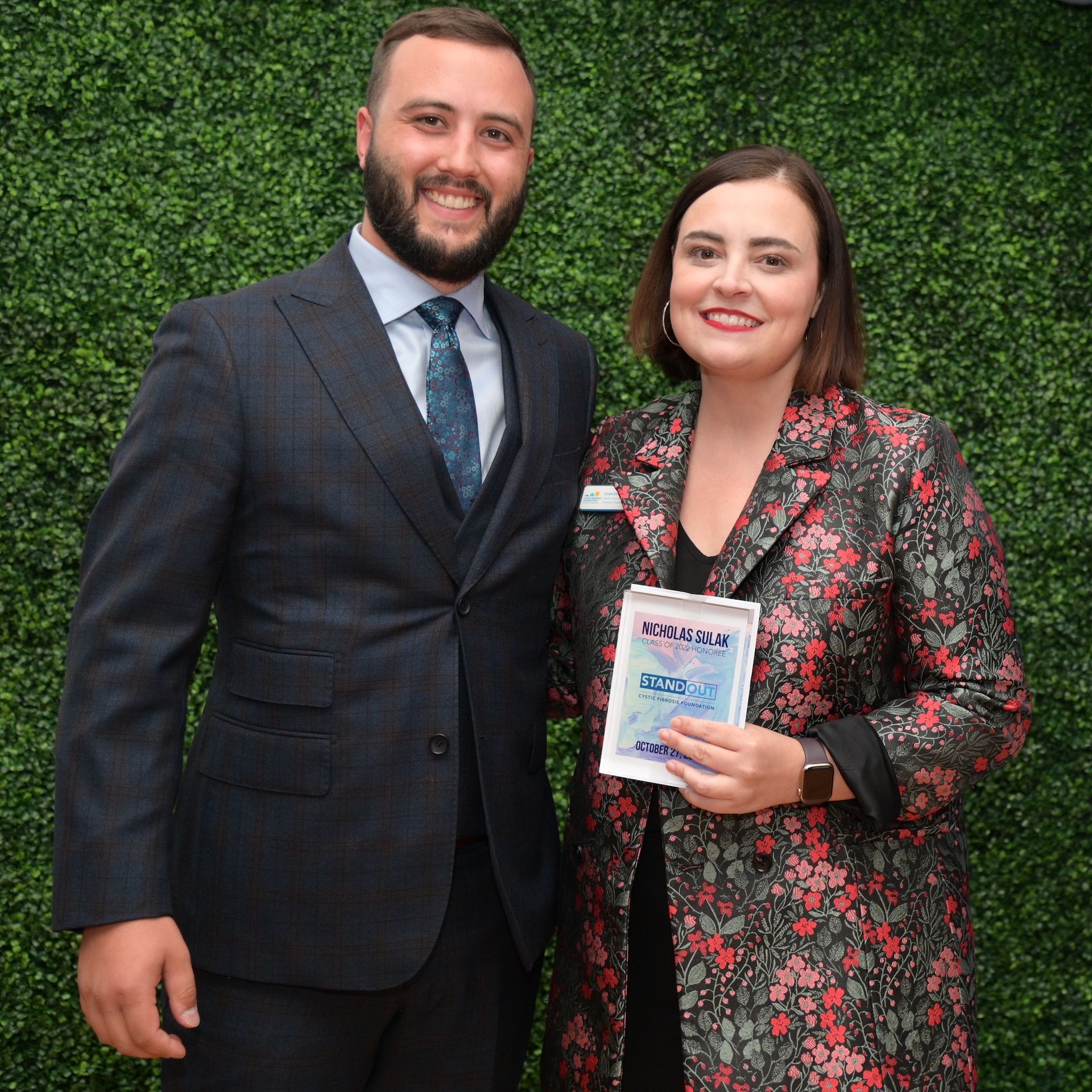Two people posing before a green hedge, smiling and holding a framed certificate.