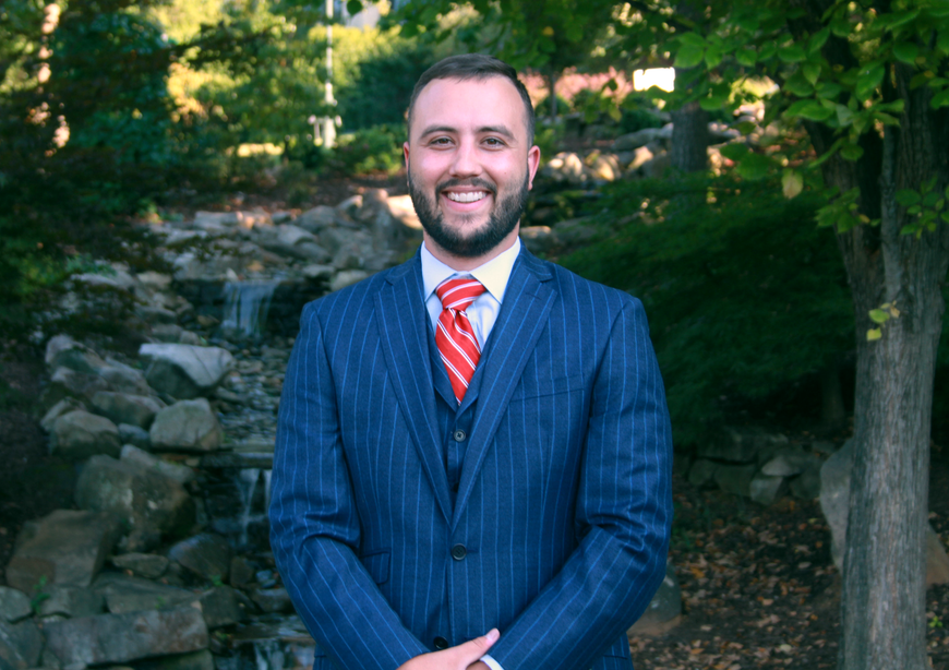 Man in blue suit and red striped tie standing outdoors in front of rocks and trees, smiling.