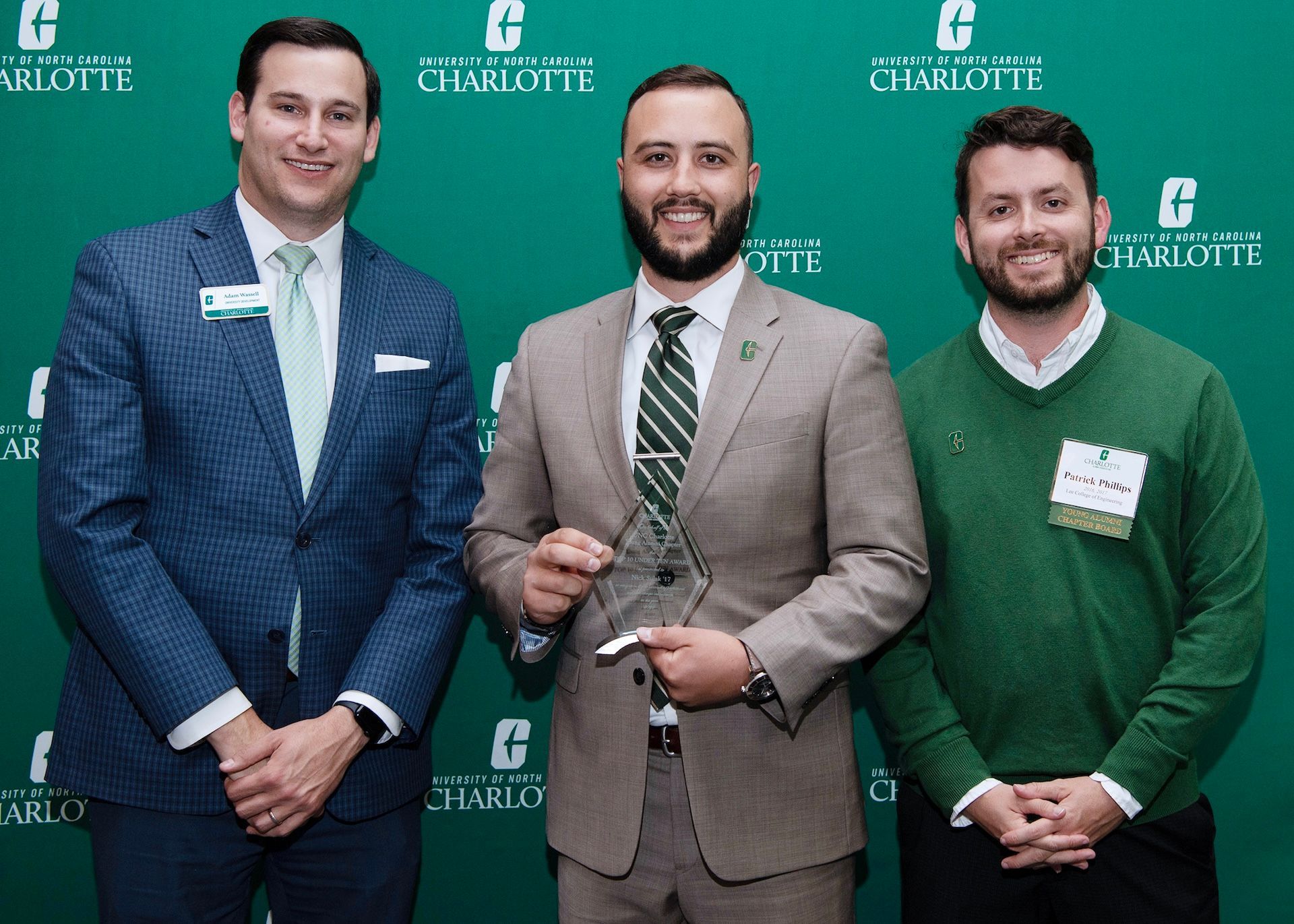 Three men posing in suits before a green Charlotte backdrop, smiling at a formal event.