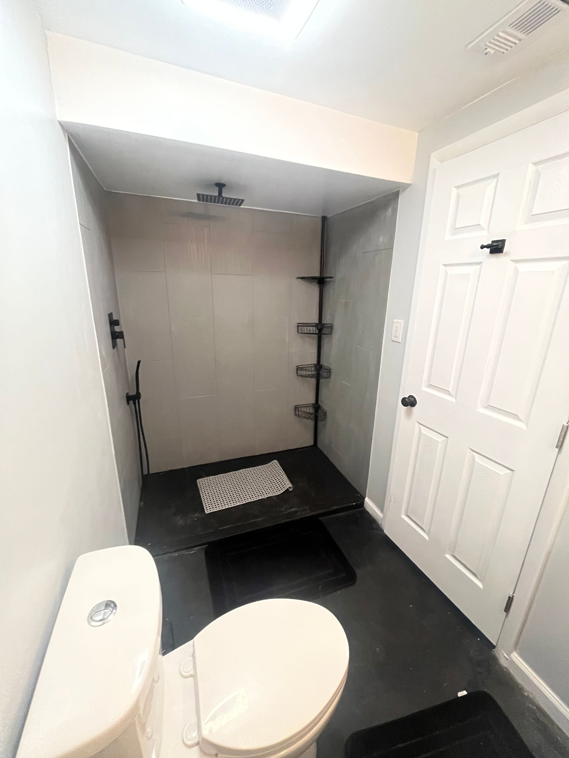 Bathroom with toilet, shower, dark floor, white door, and shower shelves.