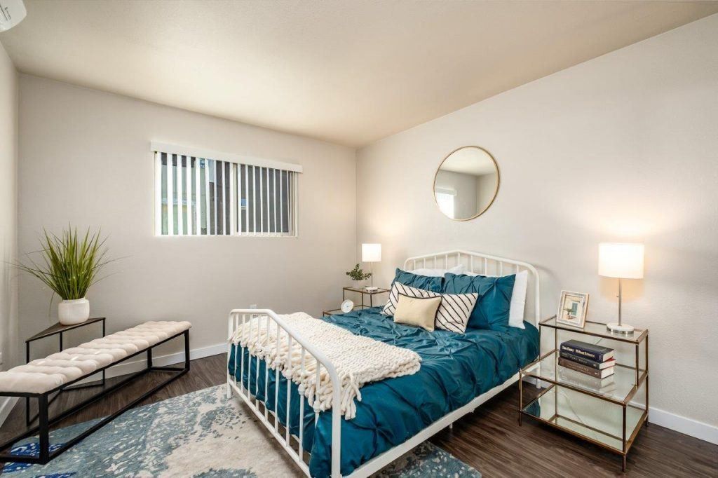 Bright bedroom with teal bedding, white walls, two bedside lamps, and a bench near the window.
