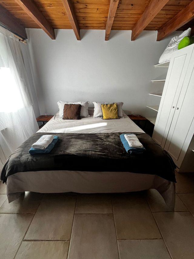 Habitación ordenada con cama hecha, armario blanco, vigas de madera a la vista y suelo de baldosas.