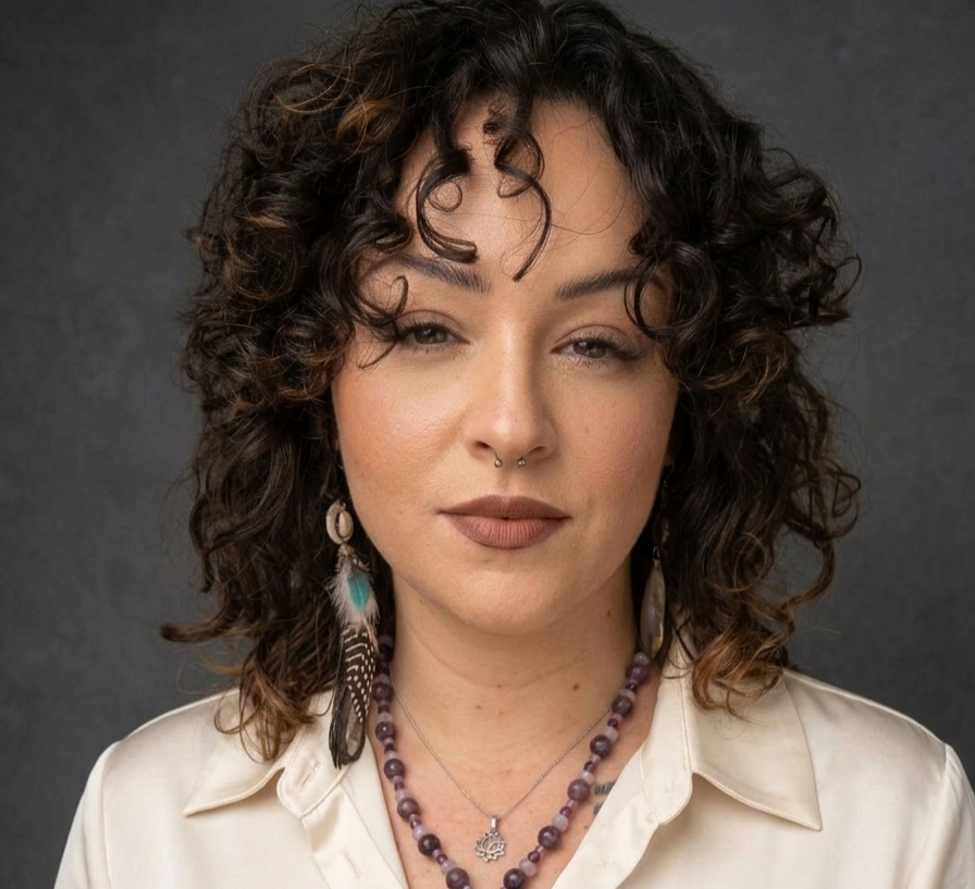 Portrait of a person with curly dark hair, wearing a white blouse and layered beaded necklaces on a gray background