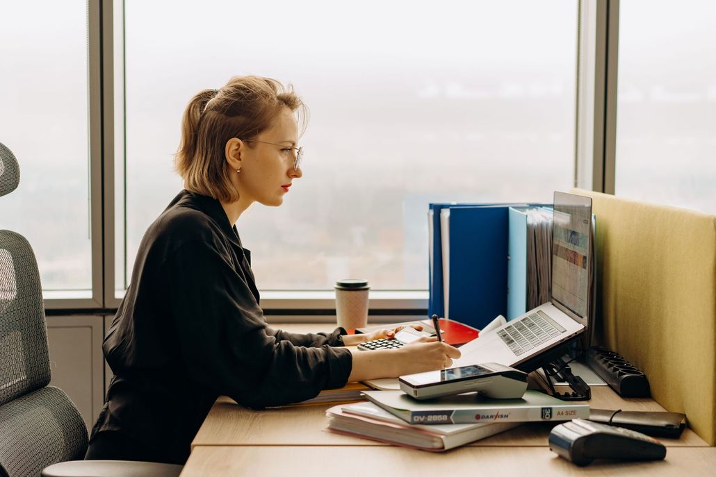 Person working at a desk with a laptop, papers, and a coffee cup in a bright office by the window