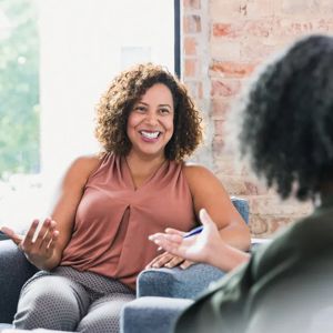 Two individuals having an engaging, seated conversation in a sunlit room with a brick wall.