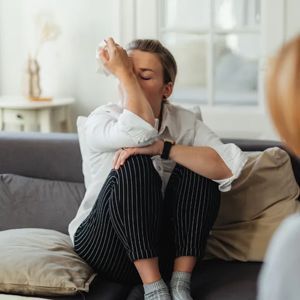A person sits on a sofa, covering their eyes with a tissue, appearing distressed during a therapy or counseling session.
