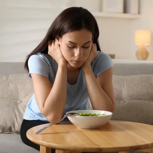 A person sits at a table with a bowl of food, looking down with a somber expression, resting their chin on their hands.