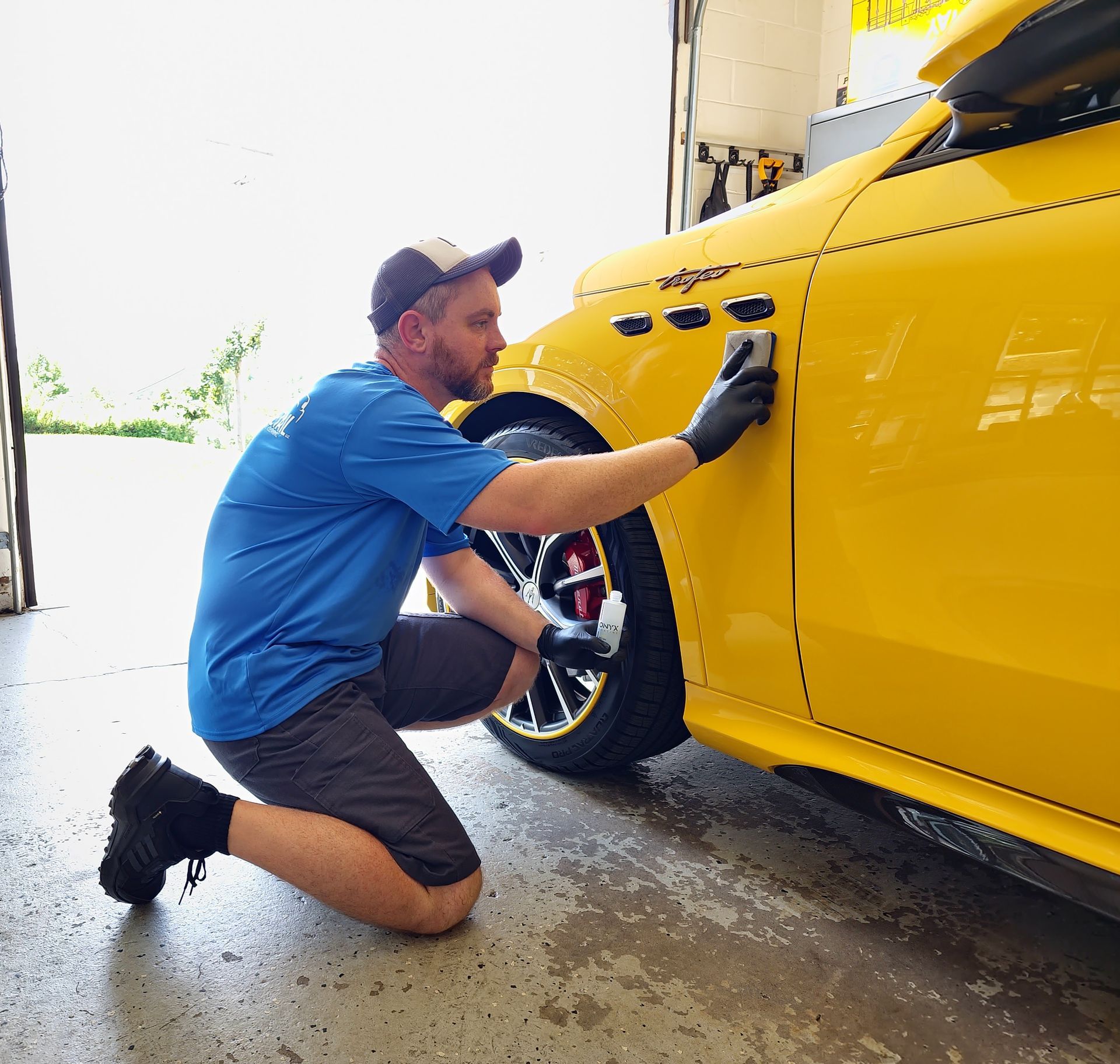 Man in blue shirt and shorts applying product to a bright yellow car, indoors.