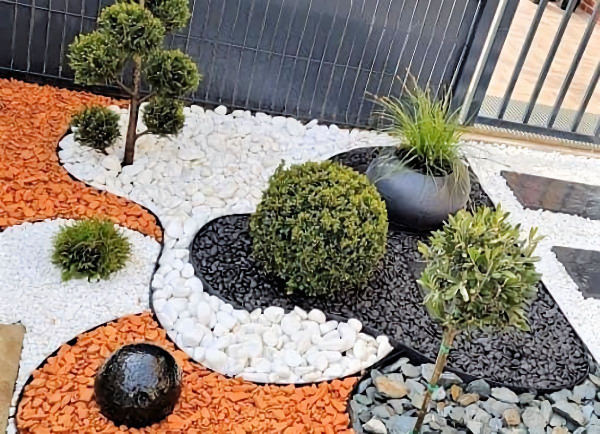 Landscaped garden bed with orange, white, and black rock surrounding trimmed shrubs and a water feature.