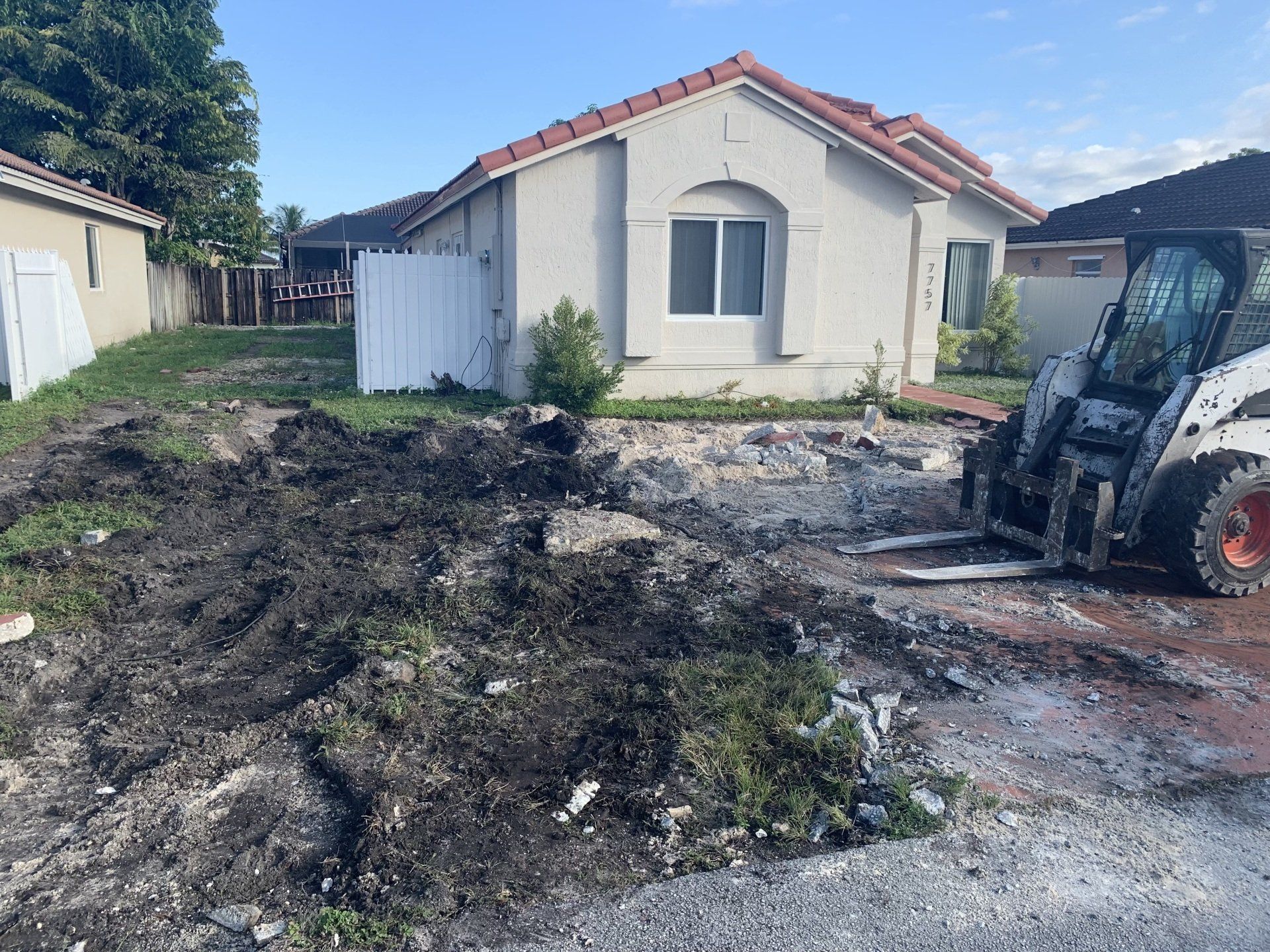 Bobcat removing dirt in front of a house. White fence and beige house. Overcast sky.