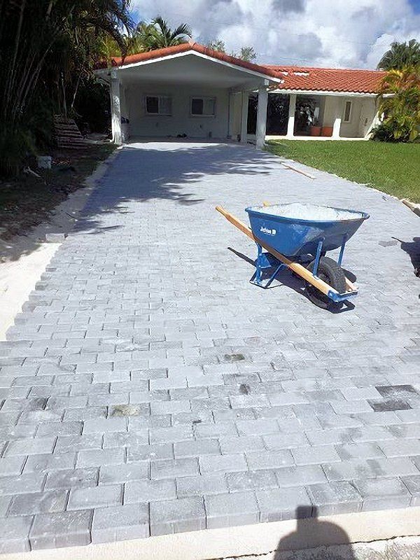 Driveway under construction with gray pavers and a blue wheelbarrow in front of a house.