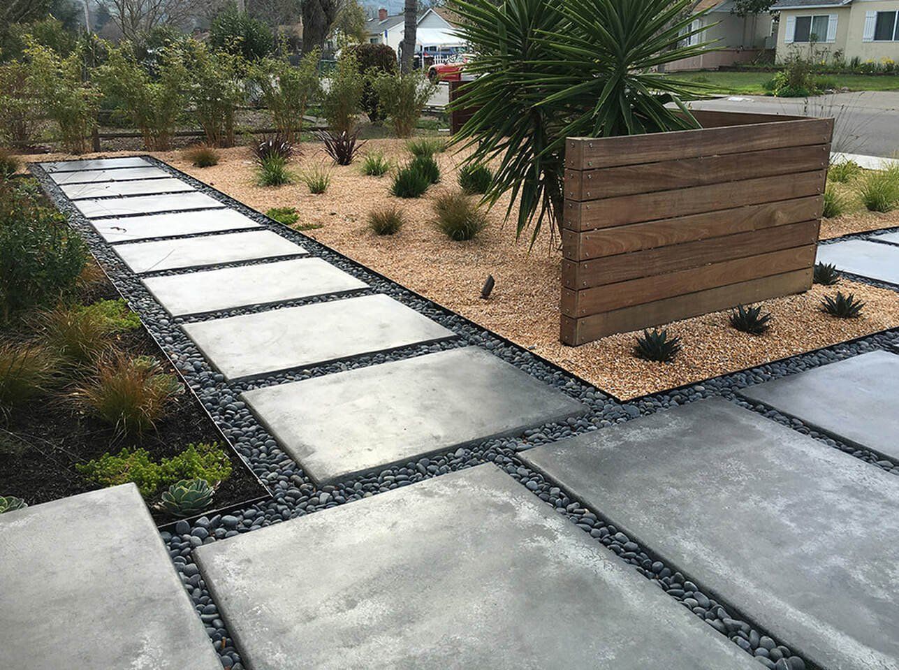 Modern walkway with large square concrete pavers, black river rocks, and wood fence.