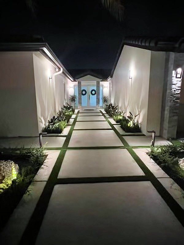 Night view of a walkway leading to double doors, lit by spotlights. Greenery lines both sides.