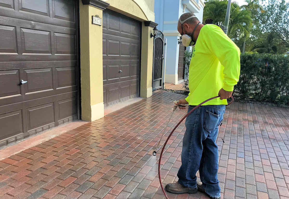 Man in safety gear power washing a paver driveway near a garage.