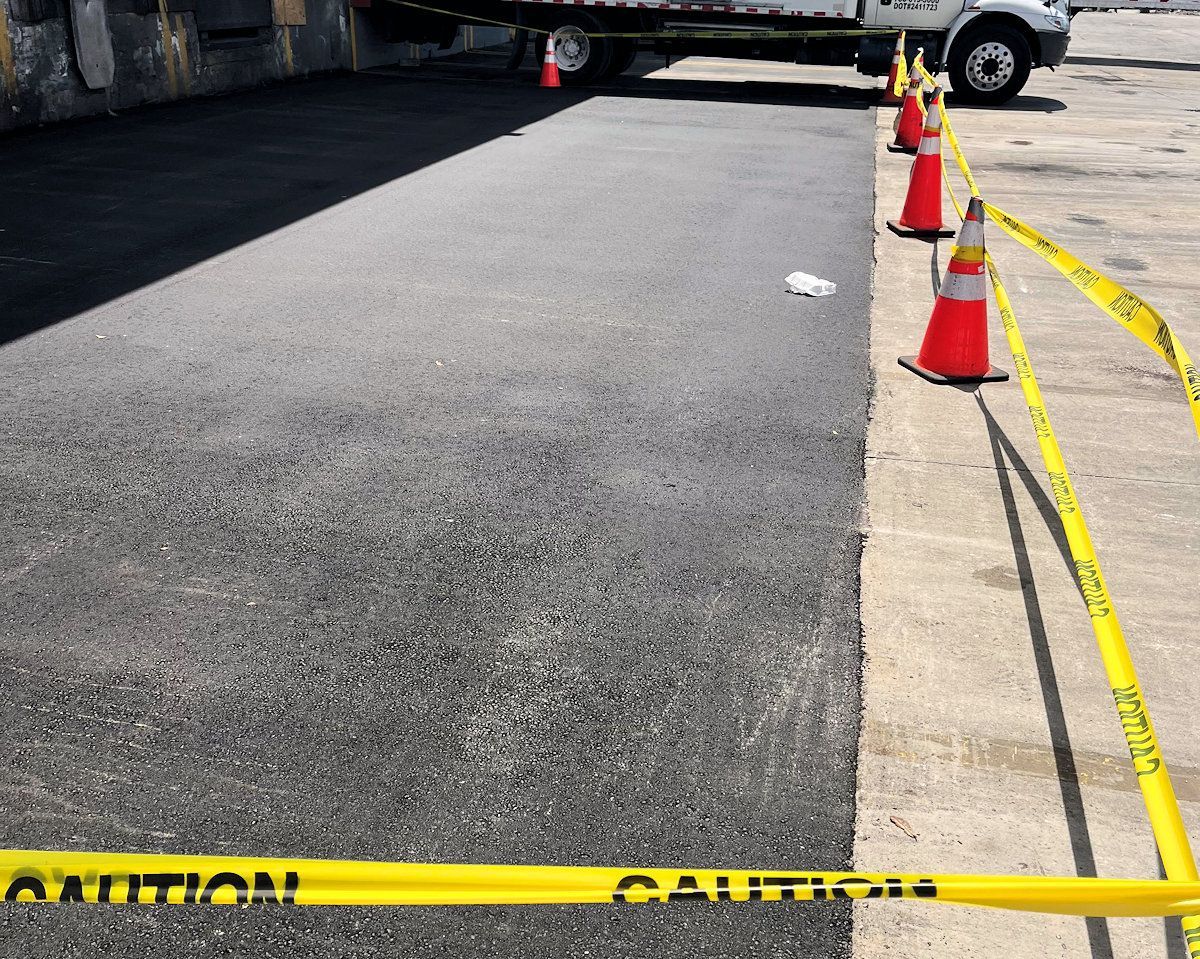 Freshly paved asphalt with caution tape and cones, next to a concrete surface.