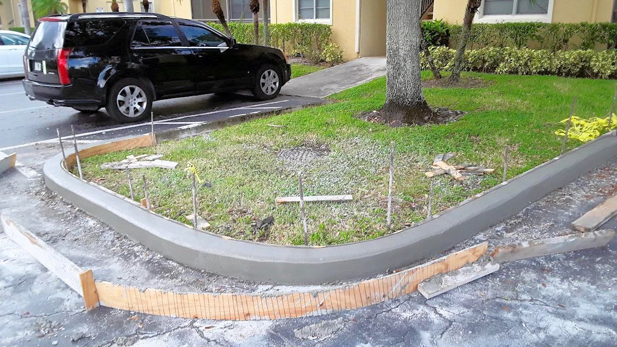 Freshly poured concrete curb along a grassy area in front of an apartment building, with a black SUV parked nearby.