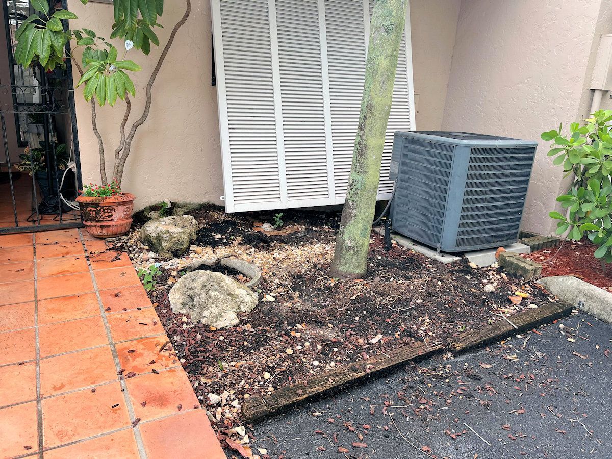 Air conditioning unit next to a tree and a covered vent in a landscaped bed with mulch and stones.