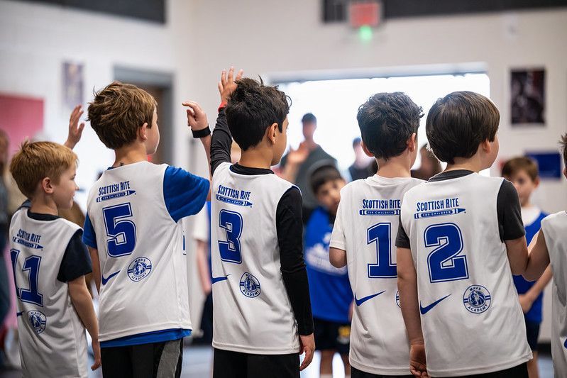 Youth volleyball team in white jerseys with blue numbers standing in a gym, raising hands together.