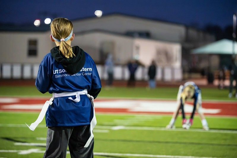 Girl in blue stands on a lit football field while another bends over in the background at dusk
