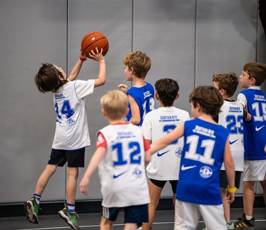 Youth basketball players in blue and white jerseys contest a shot near the hoop.