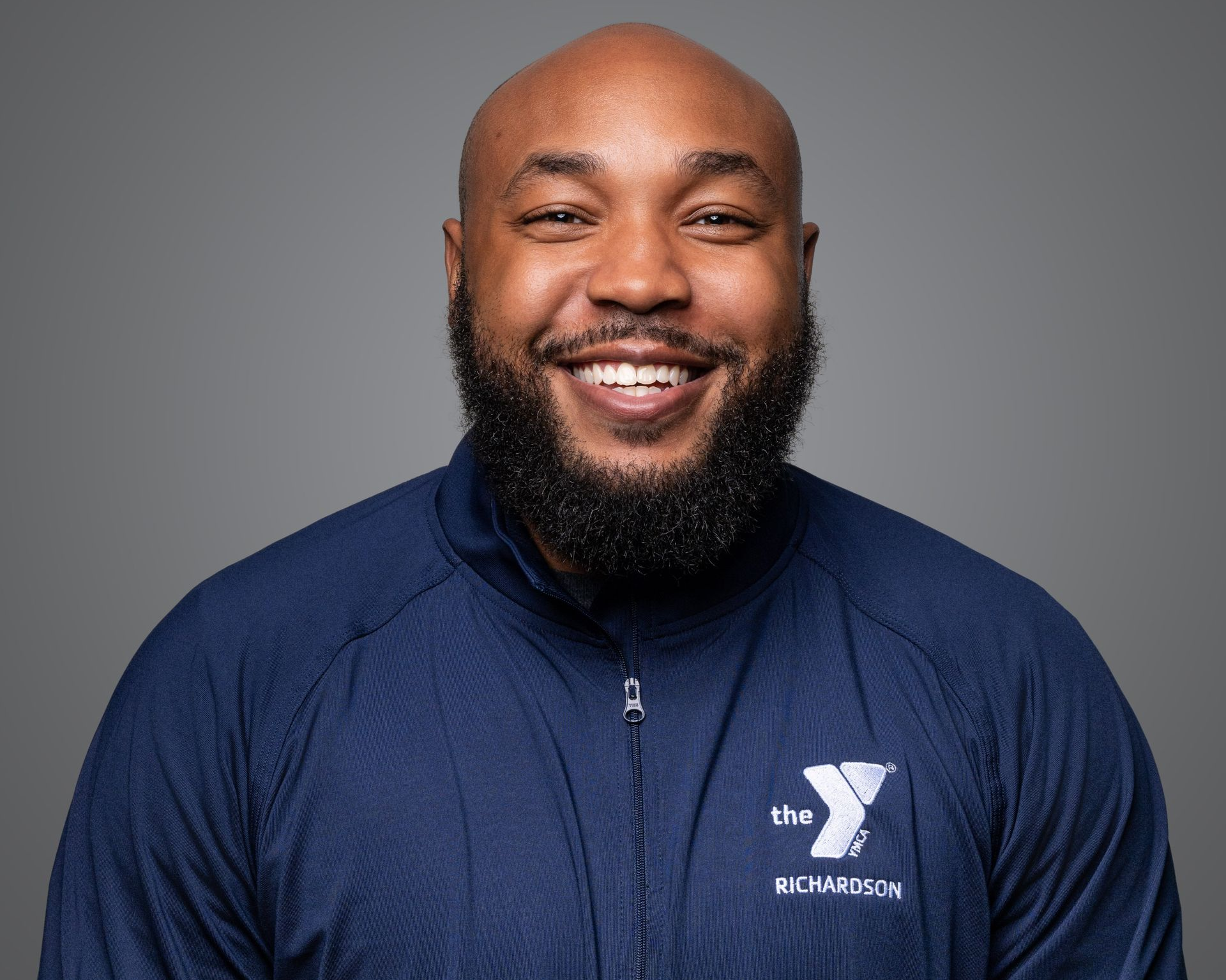 Smiling man in a navy YMCA zip-up jacket against a gray studio background