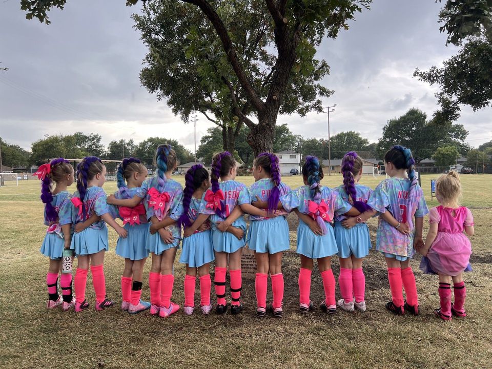 Children in blue outfits and pink socks stand in a line outdoors, holding pink pom-poms near a tree.