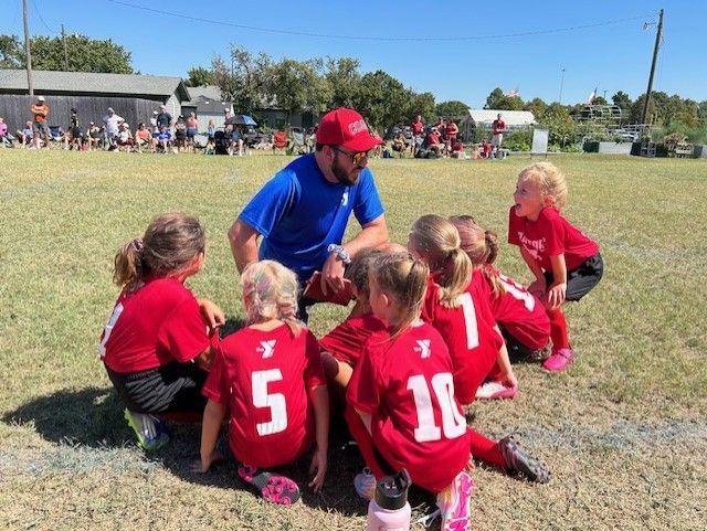 Youth soccer coach kneels with children in red jerseys on a grassy field during a game break