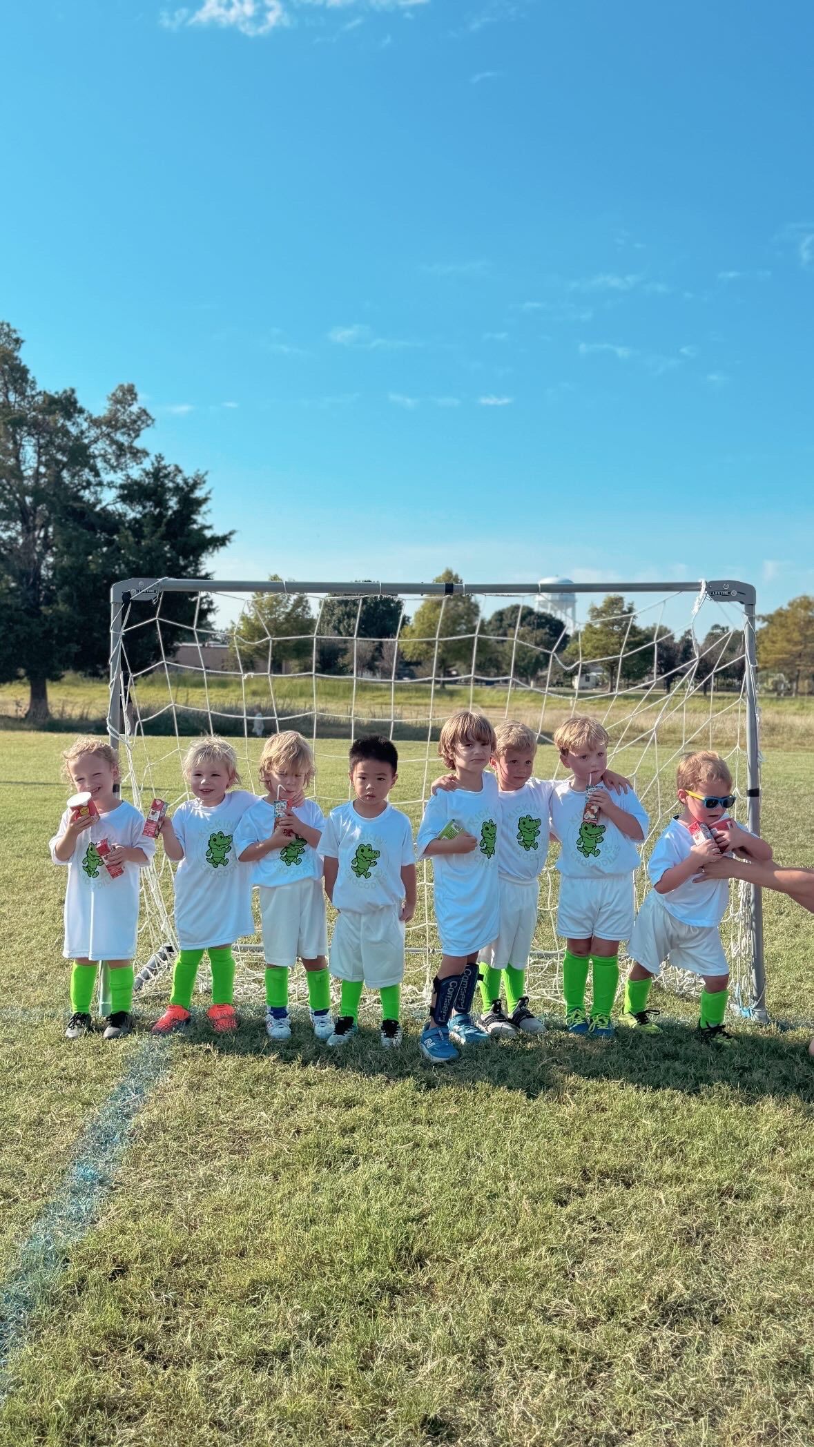 Children in white uniforms posing on a soccer field in front of a goal under a clear blue sky