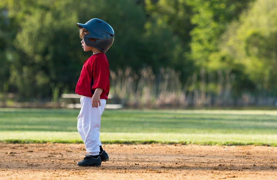 Child in a blue helmet, red shirt, and white pants standing on a baseball field.