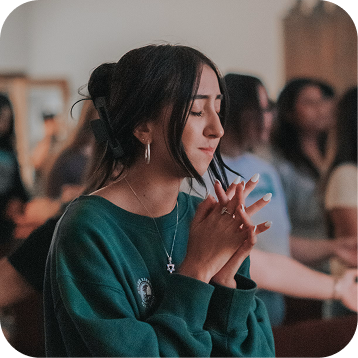 Woman with closed eyes and clasped hands, standing in a softly lit gathering