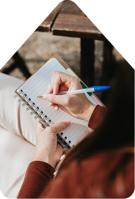 Person writing in a spiral notebook with a blue pen while seated outdoors