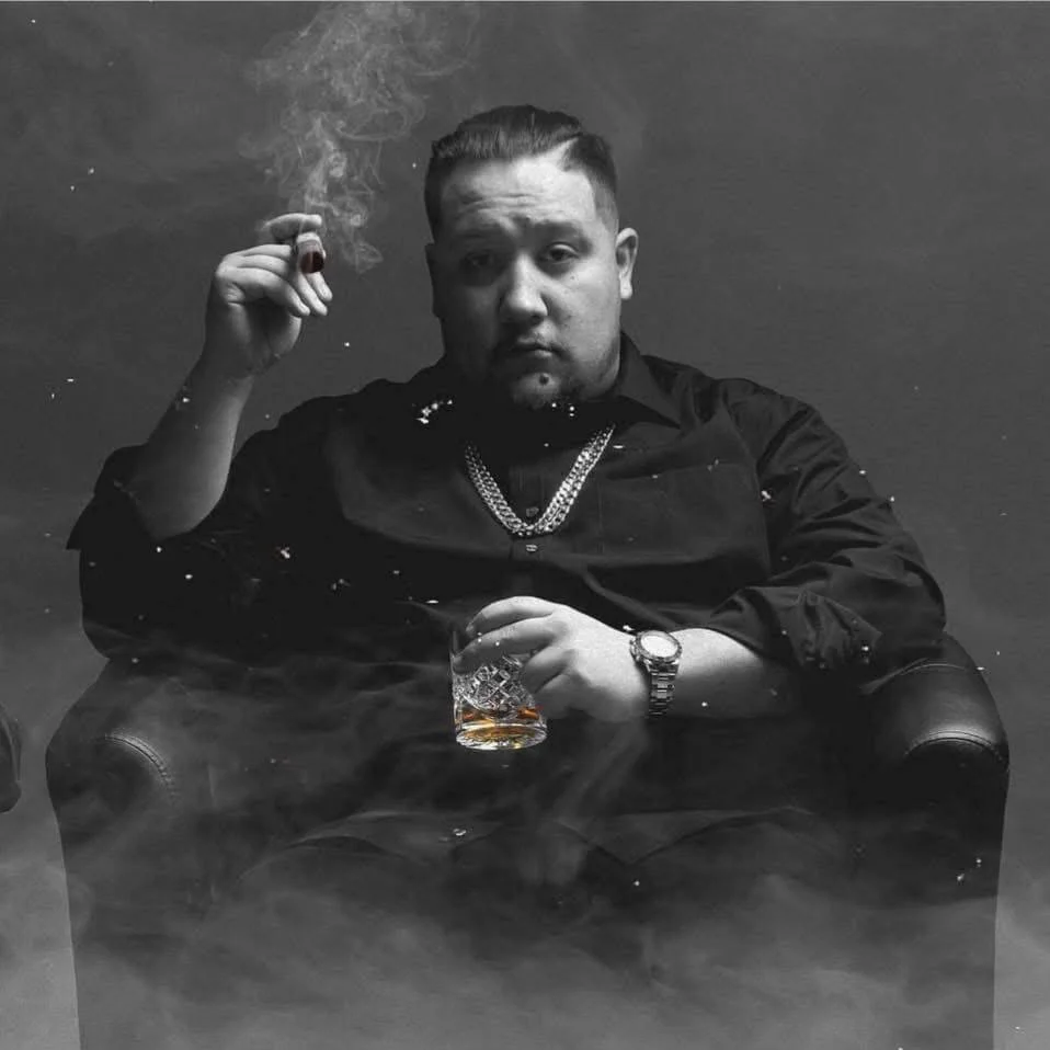 Man in black sits in a smoky room holding a drink and a cigar, wearing a chain necklace.