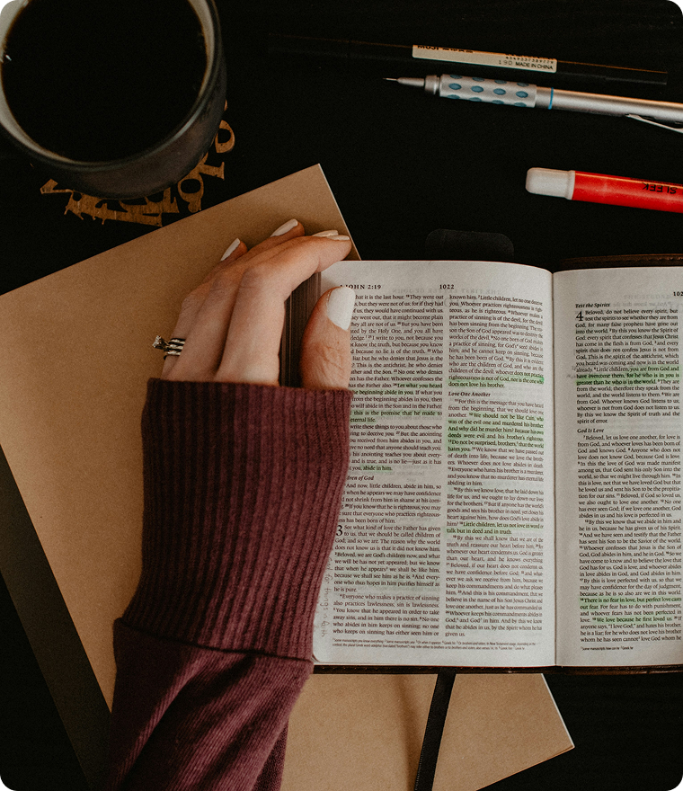 Hand holding an open book on a desk with coffee, notebook, and pens nearby