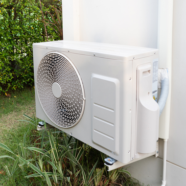 Outdoor air conditioning unit mounted beside a white house wall, with green plants nearby
