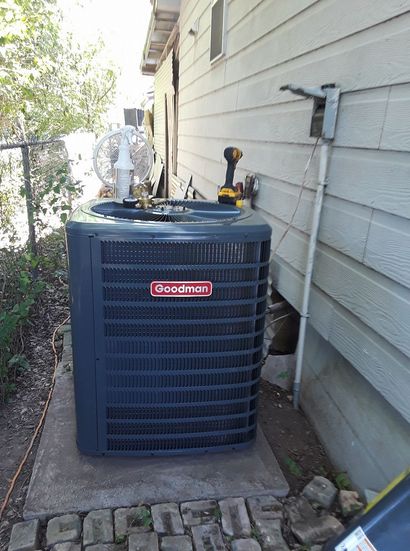 Outdoor Goodman air conditioner unit beside a house wall with a service gauge on top
