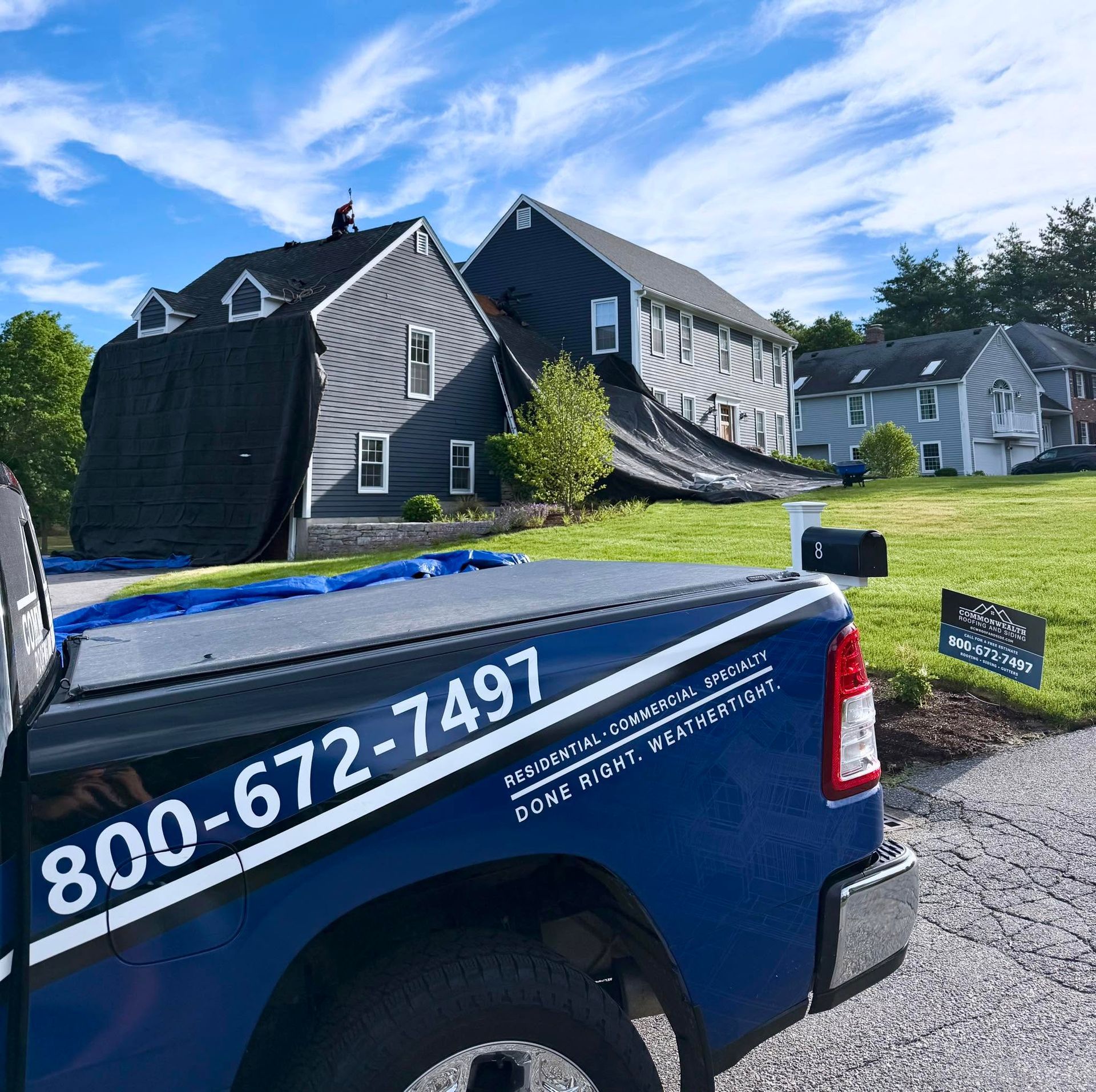 Blue pickup truck with 800-672-7491 beside storm-damaged house and roof repair tarp under a blue sky