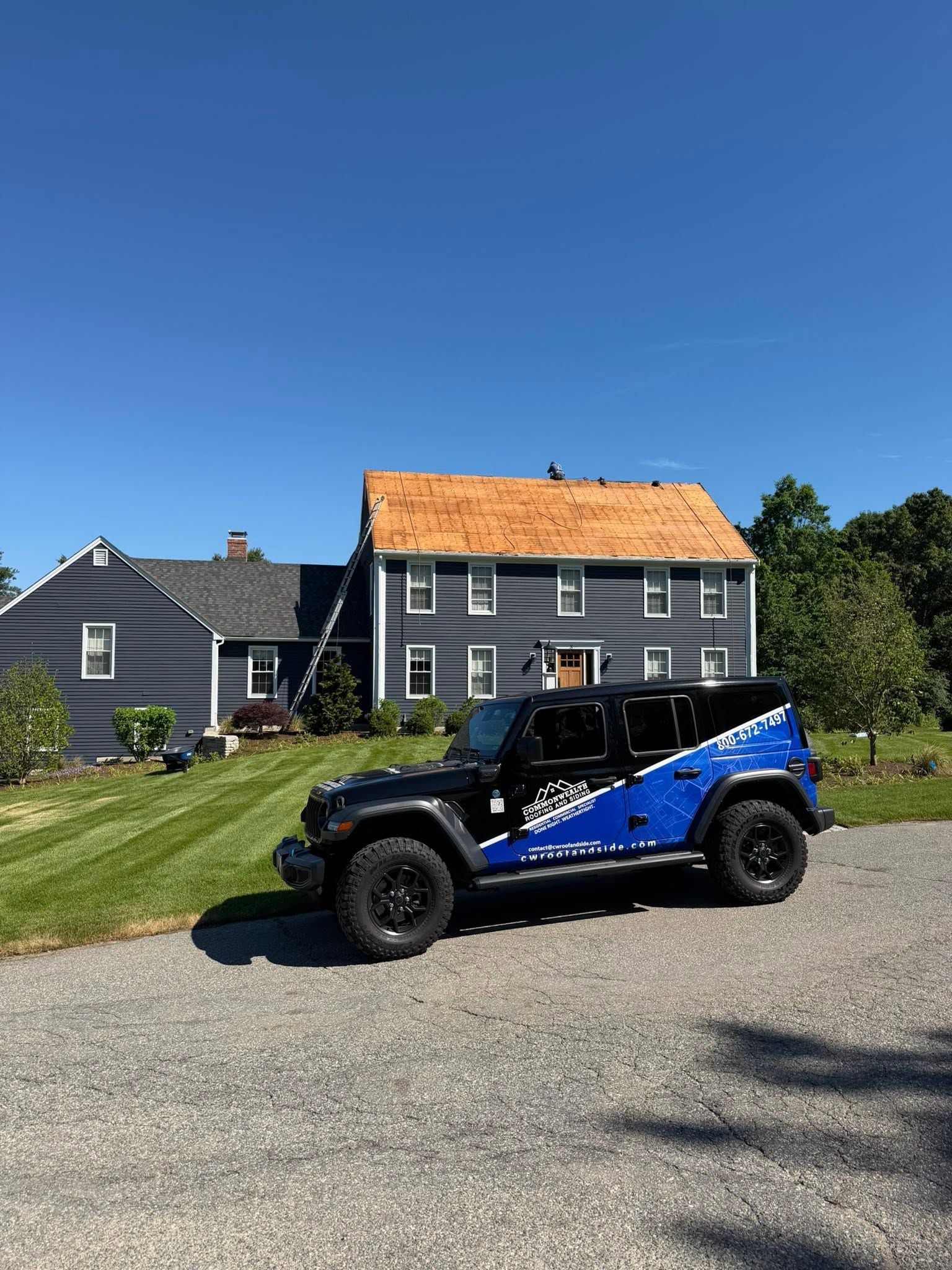 Black Jeep Wrangler parked on gravel in front of a gray house with a tan roof and blue sky.