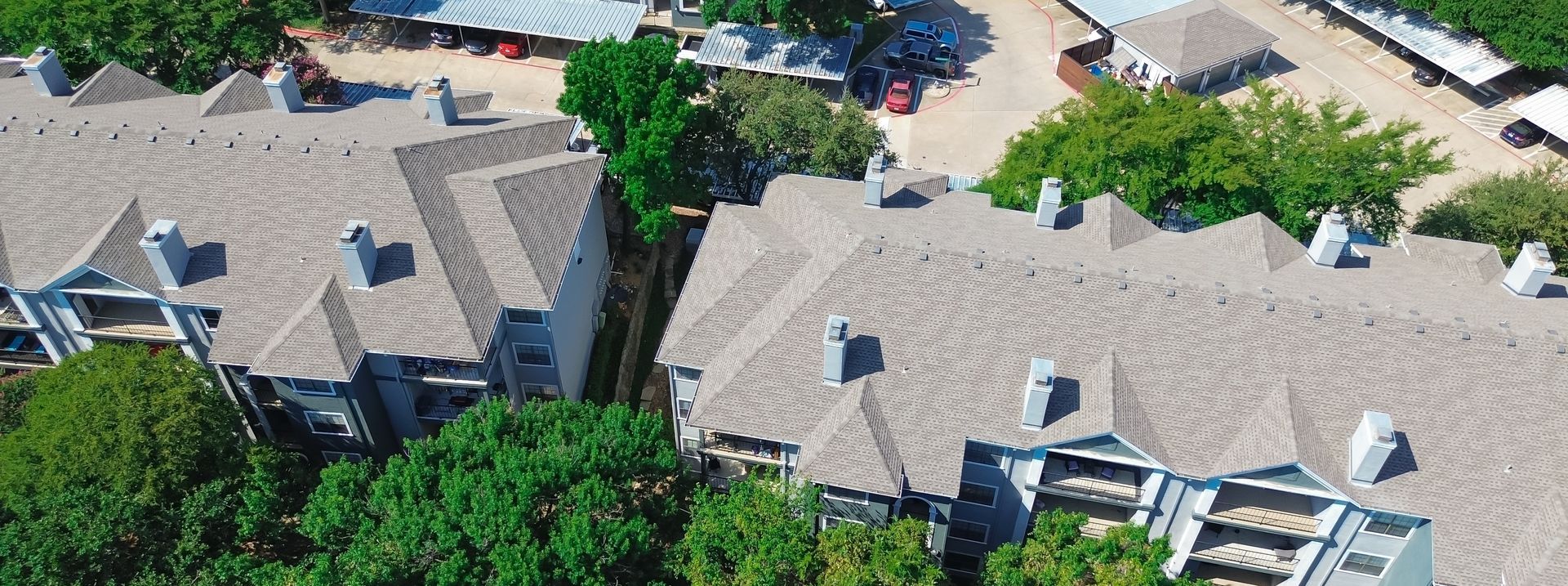 Aerial view of a gray-roofed residential complex with trees and parked cars along a street.