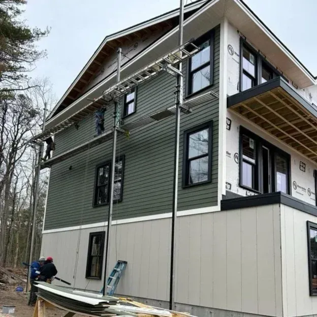 Workers on scaffolding beside a modern house under construction, with green siding and cream lower walls.