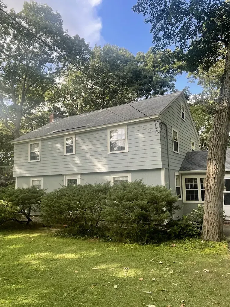 Two-story gray house with white trim, surrounded by trees and shrubs on a sunny lawn.