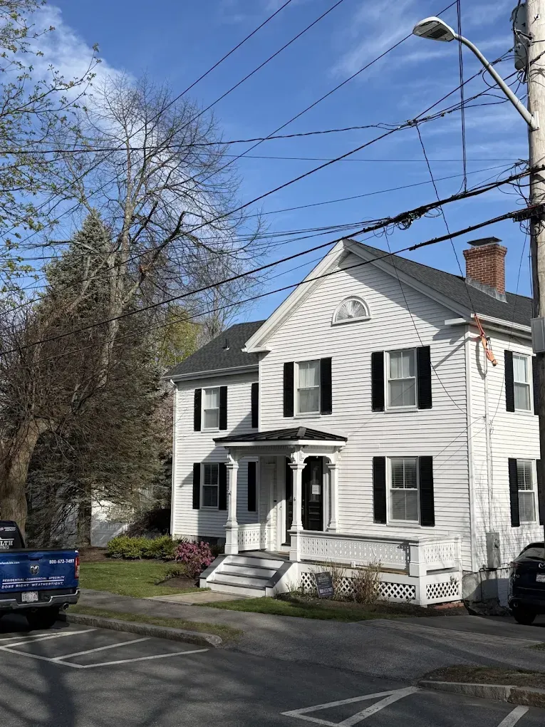 White two-story house with black shutters on a tree-lined street under a blue sky