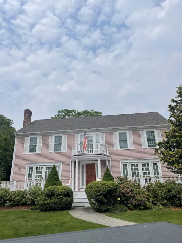 Pink colonial-style house with white shutters, a front porch, and a manicured lawn under a cloudy sky