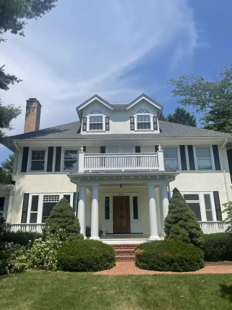 White two-story colonial-style house with balcony, columns, shutters, and a manicured front yard under a blue sky